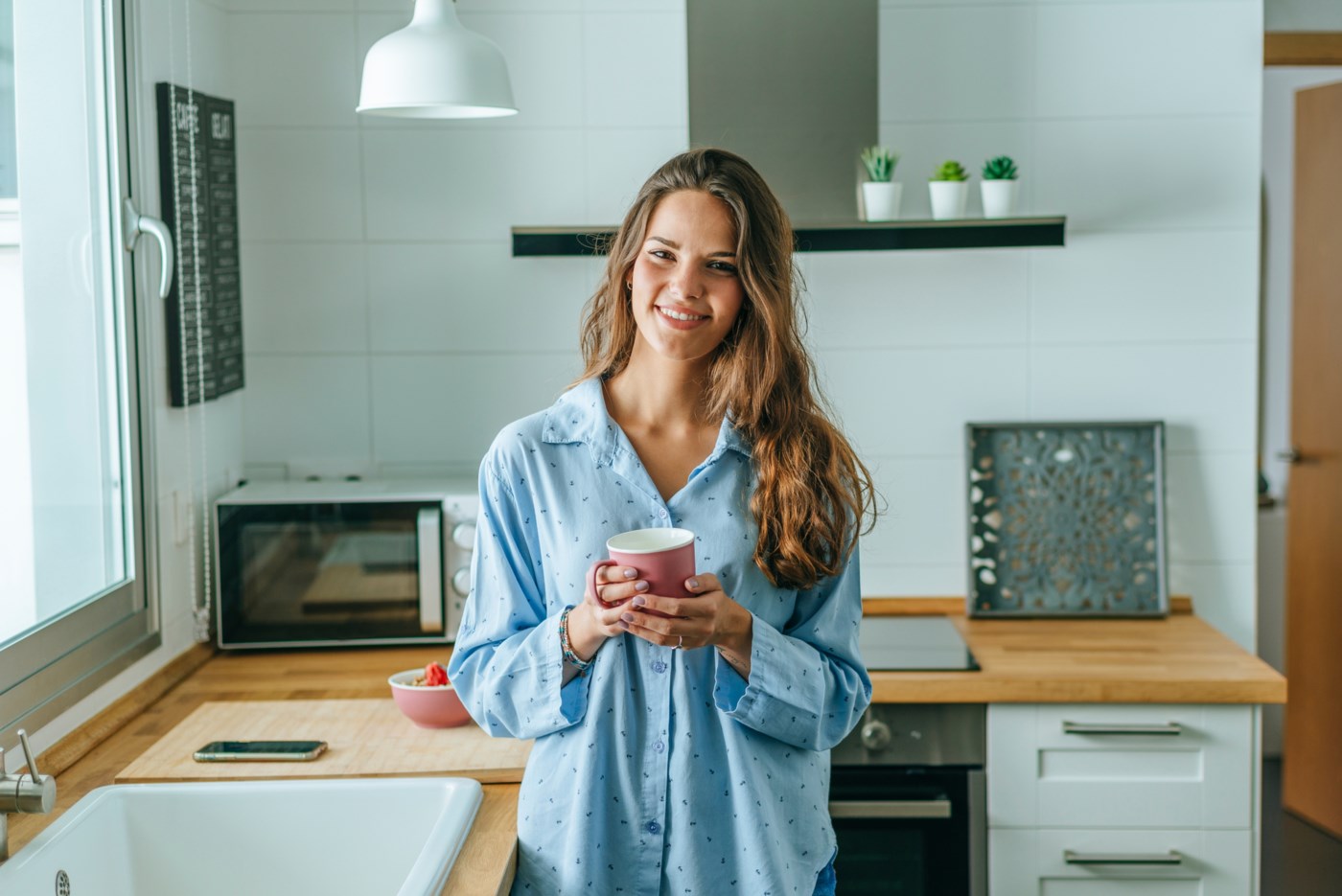 En ung kvinne smiler mot kameraet mens hun holder en kaffekopp, står på et moderne kjøkken med trebenker og hvite skap.