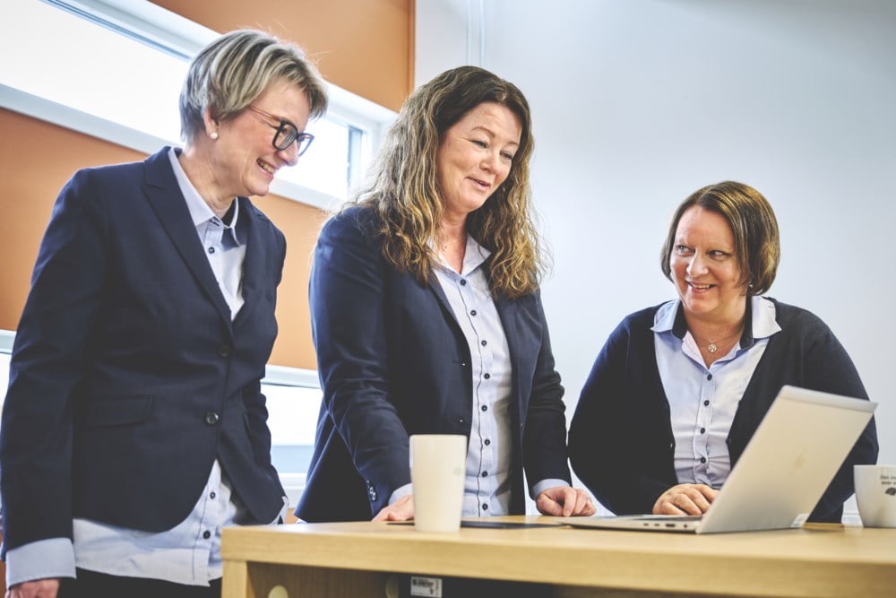 Tre blide rådgivere i Trøgstad Sparebank står ved et bord og snakker sammen. På bordet står det en laptop og kaffekopper.